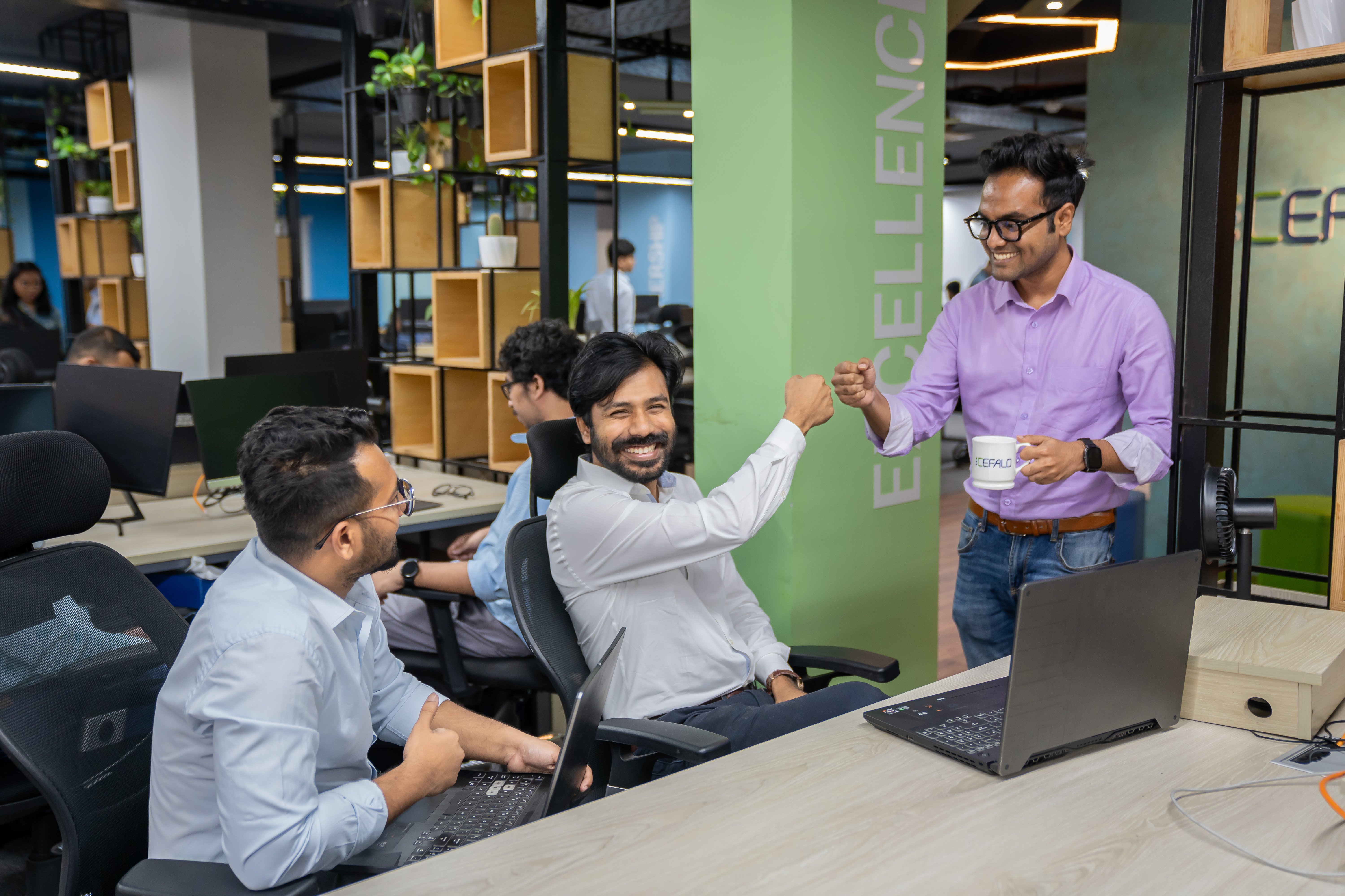 tre menn som smiler til kamera i en arbeidssetting i åpent kontorlandskap